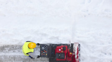 Overhead shot of a worker in hi-vis safety vest and yellow hard hat clearing snow using a snowblower.