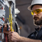 Electrical contractor working on a switchboard, wearing a hard hat and safety glasses.