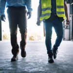 Two people walking across a warehouse, one wearing a high-visibility safety vest.